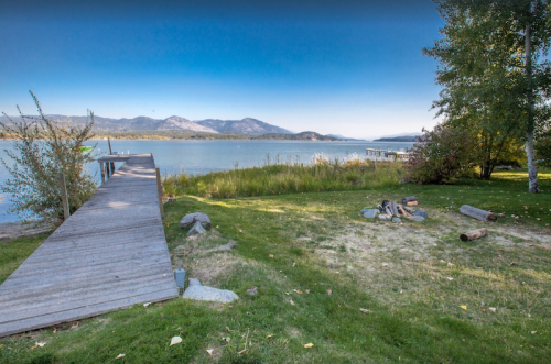 A serene lakeside view featuring a wooden dock, grassy area, and distant mountains under a clear blue sky.