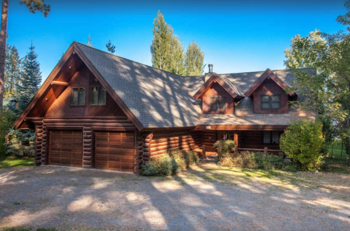 A large log cabin with a steep roof, surrounded by trees and a gravel driveway.