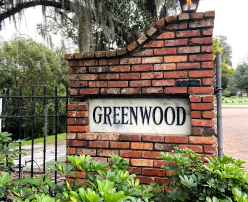 Brick entrance sign reading "GREENWOOD" surrounded by greenery and trees.