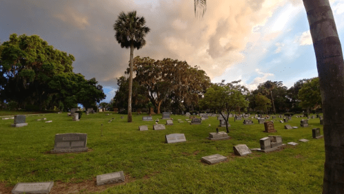 A serene cemetery scene with gravestones on green grass, surrounded by trees and a cloudy sky.