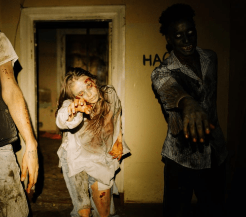 Two zombie-like figures with bloodied clothes and eerie expressions reach out in a dimly lit, abandoned room.