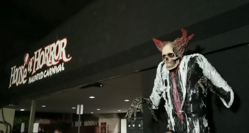 A spooky skeleton figure with red horns stands in front of a "House of Horror" sign at a haunted carnival.