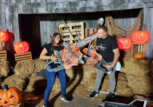 A woman and a man pose with oversized chainsaws in a spooky Halloween setting with pumpkins and hay bales.