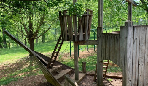 A wooden playground structure resembling a ship, with a slide, ladder, and tree canopy in a green park setting.
