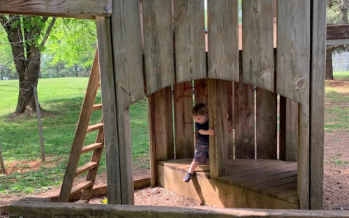 A child plays on a wooden playground structure, peeking out from behind a wooden panel. Green grass and trees in the background.
