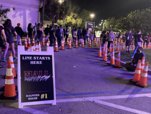 A line of people waiting for a haunted house at night, marked by traffic cones and a sign that says "Line Starts Here."