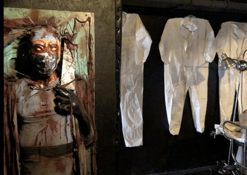 A creepy figure with glowing eyes and a mask stands beside blood-stained white outfits hanging on a dark wall.