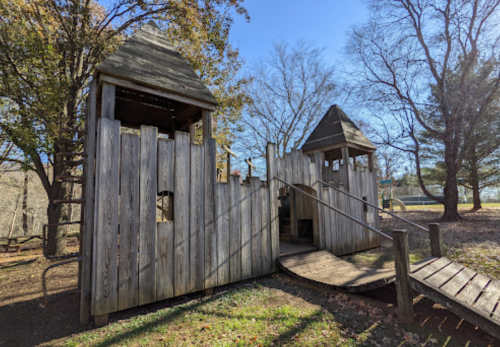 A wooden playground structure resembling a castle, with towers and a ramp, surrounded by trees and clear blue sky.