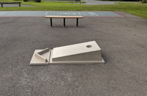 A concrete ramp with a hole, placed on a paved area near a bench and a hopscotch court in a park.