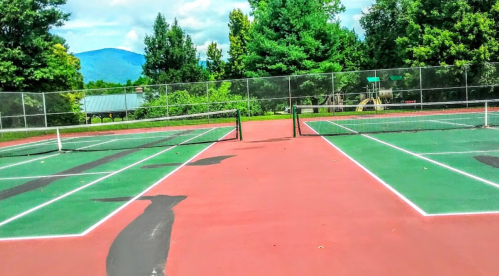 Two empty tennis courts with green and red surfaces, surrounded by trees and mountains in the background.