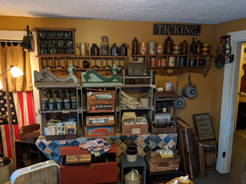 A vintage room filled with shelves of jars, boxes, and various antiques against a warm yellow wall.