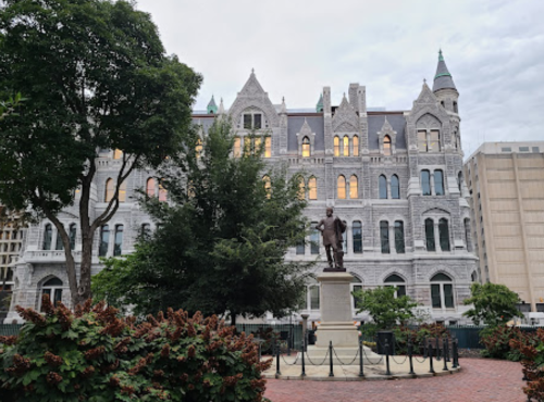 A historic building with a statue in front, surrounded by trees and shrubs, under a cloudy sky.
