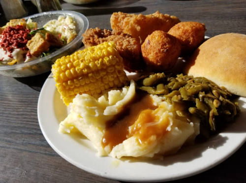 A plate of comfort food featuring mashed potatoes, corn, green beans, fried items, and a roll, with a salad in the background.