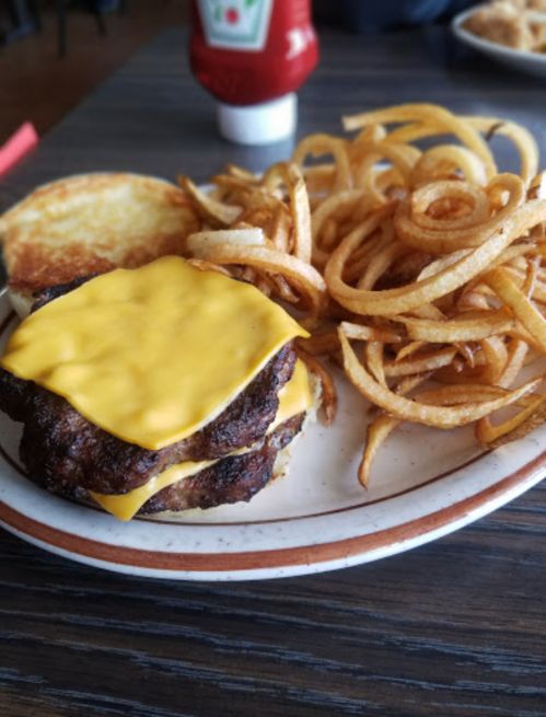 A plate with a double cheeseburger topped with melted cheese and a side of curly fries. Ketchup bottle in the background.
