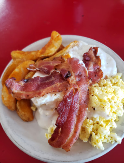 A plate of scrambled eggs, crispy bacon, and potato wedges topped with creamy gravy on a red table.