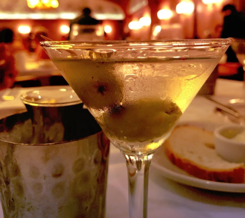 A close-up of a martini glass with olives, set on a table with a silver shaker and bread in a dimly lit restaurant.
