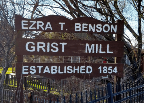 Sign for Ezra T. Benson Grist Mill, established in 1854, surrounded by trees and a fence.