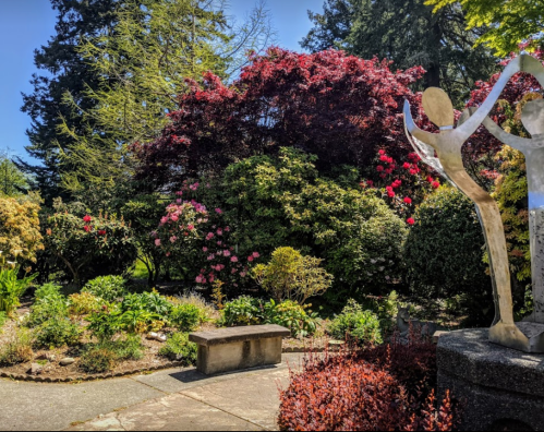 A vibrant garden with blooming flowers, lush greenery, and a modern sculpture under a clear blue sky.