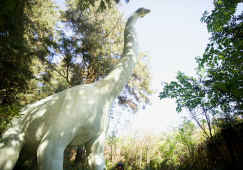 A tall dinosaur statue surrounded by trees, with a person standing near its base for scale.