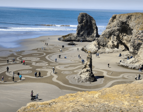A beach scene with intricate sand patterns, people walking, and rocky formations under a clear blue sky.