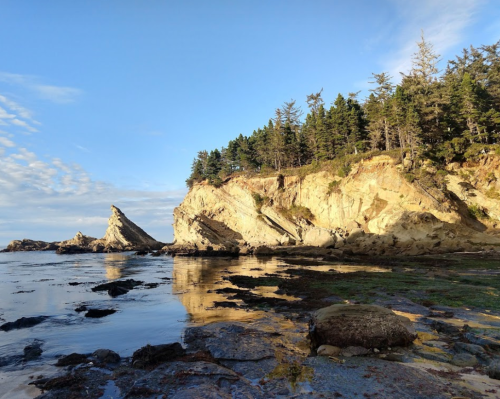 Rocky coastline with steep cliffs, lush trees, and calm water reflecting the sky under a clear blue atmosphere.