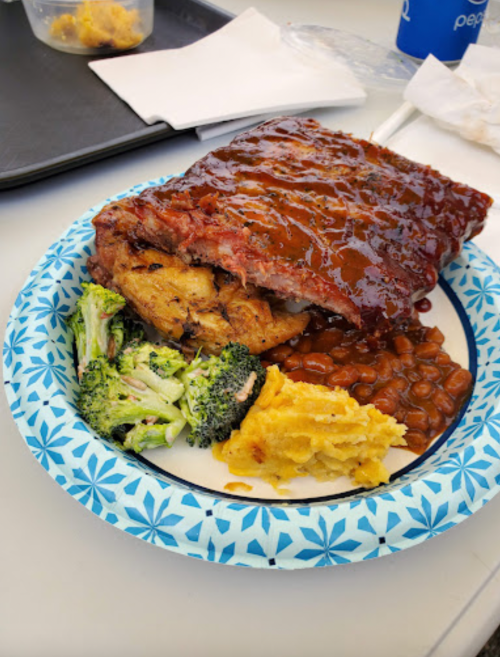 A plate with BBQ ribs, stuffing, broccoli, baked beans, and mashed potatoes on a paper plate.
