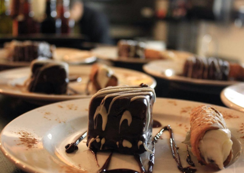Plates of chocolate desserts with drizzled sauce and a cannoli, set in a dimly lit restaurant.