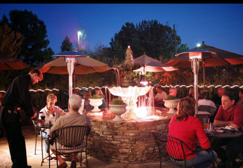 A cozy outdoor restaurant scene at night, featuring a fountain, diners at tables, and patio heaters under umbrellas.