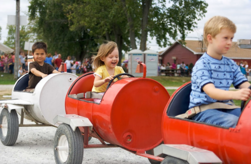 Three children ride in colorful, barrel-shaped cars on a gravel path, smiling and enjoying a fun outdoor event.