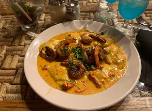 A plate of shrimp and ravioli in a creamy sauce, garnished with herbs, on a rustic table with drinks in the background.