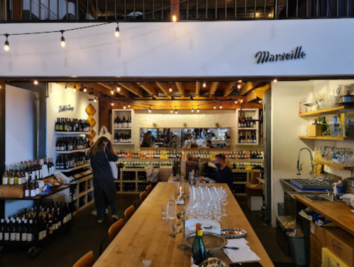 A cozy wine shop with shelves of bottles, a tasting area, and warm lighting, featuring people browsing and preparing for a tasting.