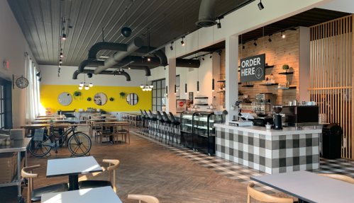 A modern café interior with a yellow accent wall, seating areas, and a counter displaying pastries and drinks.