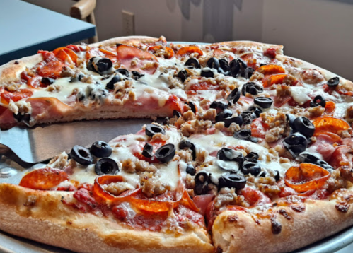 A close-up of a pizza topped with pepperoni, sausage, black olives, and melted cheese, with a slice removed.