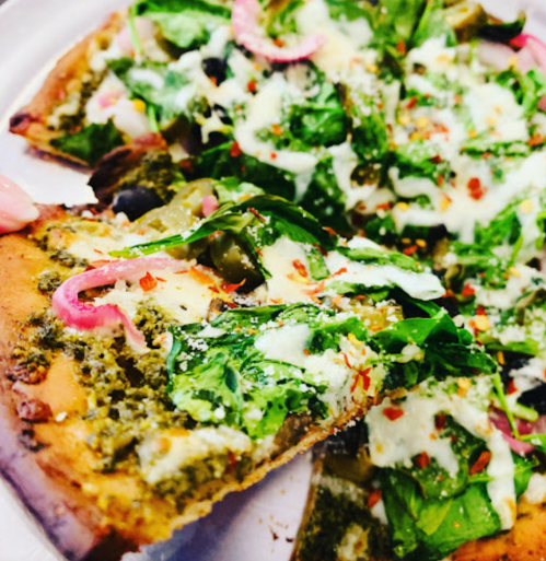 A close-up of a slice of pesto pizza topped with spinach, olives, red onions, and cheese, garnished with red pepper flakes.