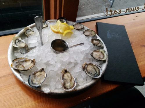 A metal platter of fresh oysters on ice, garnished with lemon slices and served with a small dipping sauce.