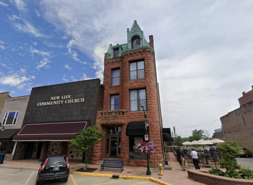 Historic red stone building with a green turret, housing New Life Community Church, surrounded by trees and outdoor seating.