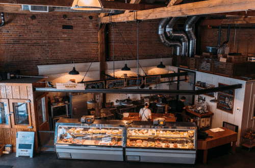 A cozy butcher shop with a display case of meats, wooden beams, and staff preparing food in a warm, inviting atmosphere.