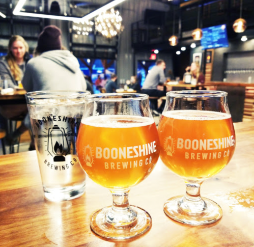 Two glasses of amber beer with the Booneshine Brewing Co. logo, set on a wooden table in a lively brewery.