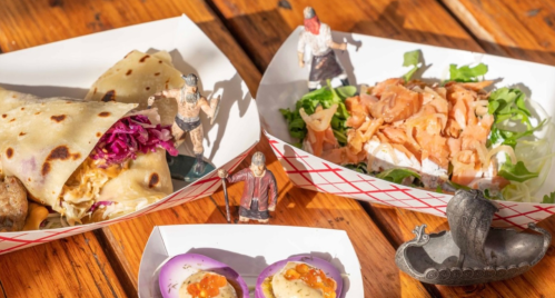 A colorful spread of food featuring a wrap, salad, and small dishes, accompanied by toy figures and a decorative boat.