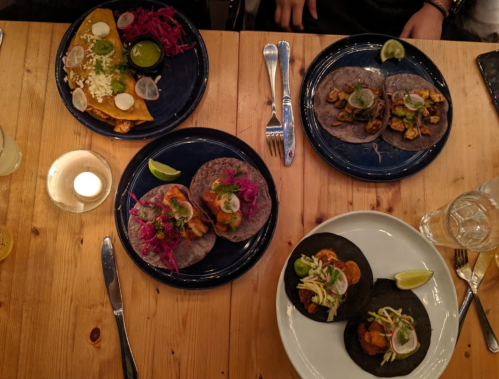 A wooden table with four plates of colorful tacos, garnished with lime and vegetables, alongside a candle and drinks.