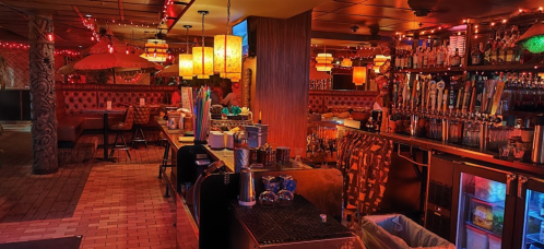 A dimly lit bar with tiki decor, colorful lights, and a variety of drinks on tap, creating a vibrant atmosphere.