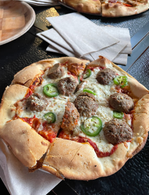 A close-up of a pizza topped with meatballs, jalapeños, and melted cheese, served on a textured black table.