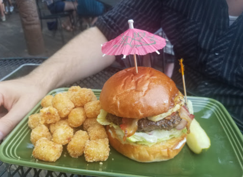 A burger with bacon, lettuce, and an umbrella pick, served with crispy tater tots on a green plate.