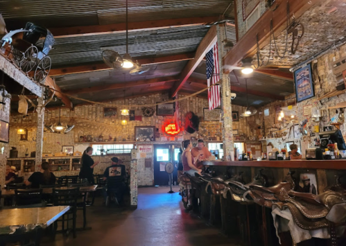 A rustic restaurant interior with wooden beams, brick walls, and patrons at the bar, decorated with memorabilia and an American flag.