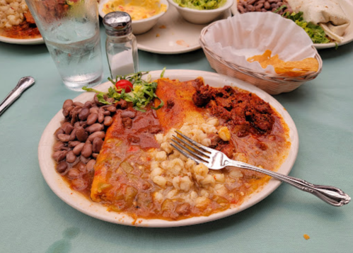 A plate of enchiladas topped with sauce, served with beans, corn, and a side salad on a table with drinks.