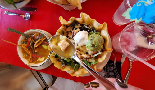 A hand holds a spoon above a colorful taco salad in a crispy bowl, with a side of salsa and garnishes on a red table.