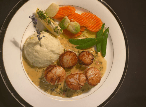 A gourmet plate featuring seared scallops, creamy mashed potatoes, and colorful vegetables in a rich sauce.