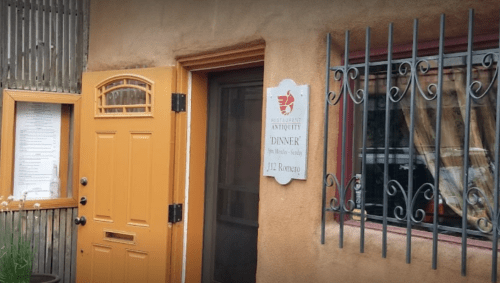 Exterior view of a restaurant entrance with a yellow door and a sign reading "Antiquity." Window with decorative curtains visible.