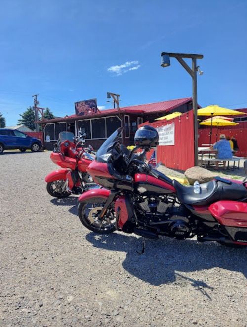 Two red motorcycles parked in front of a rustic building with outdoor seating under a clear blue sky.