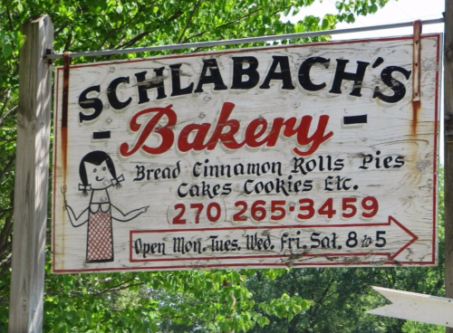 Sign for Schlabach's Bakery featuring baked goods and contact information, with an illustration of a woman. Open hours listed.